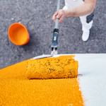 Ein Handwerker in weißer Kleidung trägt mit einem Farbroller orangene Farbe auf eine weiße Wand auf.