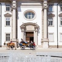 Springbrunnen und vorbeifahrende Pferdekutsche vor dem Eingangsbereich der Kirche