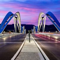 Brücke mit geschwungenen, weißen Bögen, die sich über eine Straße erstreckt. Der Himmel ist in sanften Lila- und Rosatönen gefärbt.