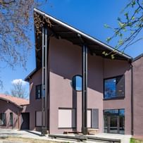 Gebäudes mit schrägen Dächern und großen Fenstern, umgeben von Bäumen und einem klaren blauen Himmel.