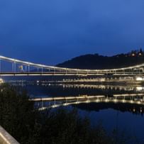 Eine nächtliche Luftaufnahme einer Brücke, die über einen Fluss führt. Die Brücke ist beleuchtet und reflektiert im Wasser.