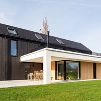 Ein zweigeschossiges Haus mit einer Kombination aus schwarzer Holzverkleidung und hellen Fassadenelementen. Die Vorderseite des Hauses hat große Fenster und eine überdachte Terrasse, die zu einem gepflegten Garten mit grünem Rasen führt. Im Hintergrund sind einige Bäume und ein klarer Himmel zu sehen.