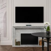 Wohnbereich einer Suite im „The Amauris“ Wien mit Kaminfront aus hellem Marmor, darüber ein Flachbildfernseher, runder Tisch mit zwei Stühlen und weißer Orchidee sowie Blick in einen angrenzenden Raum mit Sessel und bodenlangen Vorhängen auf dunklem Fischgrätparkett.