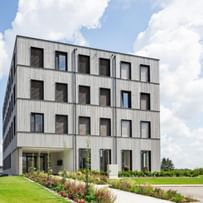 Ein mehrstöckiges Gebäude mit einer Fassade aus Holz und großen Fenstern. Es hat eine rechteckige Form und ist von einem gepflegten Garten mit Blumen und Sträuchern umgeben
