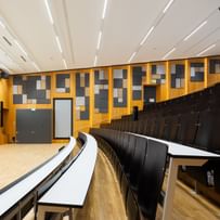 Leerer Hörsaal mit gestuften Sitzreihen und langen Tischen, grüner Tafel, doppelflügeliger Tür und Uhr. Die Wände sind mit Akustikpaneelen in verschiedenen Formen versehen, die Decke mit mehreren Lichtquellen ausgestattet.