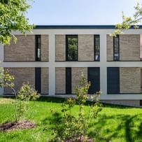 Modernes zweistöckiges Gebäude mit großen Fenstern und Balkonen, gelegen in einer grünen Umgebung mit Bäumen im Hintergrund bei sonnigem Wetter und klarem Himmel.