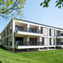 Modernes zweistöckiges Gebäude mit großen Fenstern und Balkonen, gelegen in einer grünen Umgebung mit Bäumen im Hintergrund bei sonnigem Wetter und klarem Himmel.