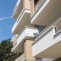 Wohnanlage in der Robert-Stolz-Straße in Linz. Ein modernes, mehrstöckiges Wohngebäude mit hellen Fassaden und zahlreichen Balkonen. Große Fenster sorgen für viel Tageslicht. Vor dem Gebäude stehen Autos auf einer ruhig wirkenden Straße, eingerahmt von gepflegten Grünflächen und Bäumen.