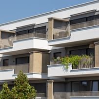 Wohnanlage in der Robert-Stolz-Straße in Linz. Ein modernes, mehrstöckiges Wohngebäude mit hellen Fassaden und zahlreichen Balkonen. Große Fenster sorgen für viel Tageslicht. Vor dem Gebäude stehen Autos auf einer ruhig wirkenden Straße, eingerahmt von gepflegten Grünflächen und Bäumen.