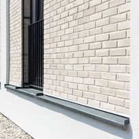  Nahaufnahme einer Gebäudefassade mit hellen Ziegeln, schwarzen Fensterbänken und Metallgittern an den Fenstern. Unten und rechts ist weißer Putz zu sehen, entlang der Wand verläuft ein Streifen mit weißen Kieselsteinen.