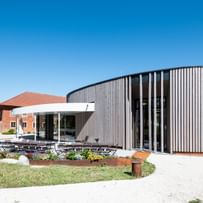 Gebäude mit einer geschwungenen Holzfassade und großen Fenstern, umgeben von einem gepflegten Garten und einer Terrasse