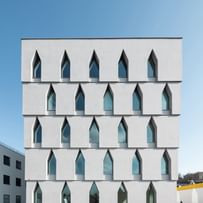 Frontale Außenansicht eines mehrstöckigen weißen Gebäudes mit großen, schmalen Fenstern, die in einer einzigartigen, spitz zulaufenden Form angeordnet sind
