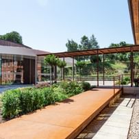 Heller, sonniger Außenbereich mit einem Weg, der zu einem Gebäude führt. Auf der linken Seite ist ein überdachter Bereich mit einer Holzstruktur zu sehen.