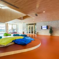 Heller Raum mit einem geschwungenen, orangefarbenen Boden. In der Mitte des Raumes befinden sich große, bunte Sitzkissen in verschiedenen Farben (grün, blau, gelb), die auf dem Boden verteilt sind. 