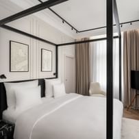 Helles Executive-Suite-Schlafzimmer im „The Amauris“ Wien mit modernem Himmelbett in schwarzem Rahmen, weißer Bettwäsche, abstrakten Wandbildern, bodentiefen Vorhängen, Lounge-Sessel und freistehendem TV auf hellem Parkett.
