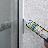 Eine Dichtmasse wird mit einer Kartuschenpistole auf eine Fensterrahmennaht aufgetragen. Die Wand ist hell und strukturiert, und die Dichtmasse wird gleichmäßig entlang der Kante aufgetragen. 