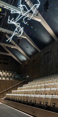 Auditorium mit einer Holzdecke und einer Sitzanordnung aus hellen, gepolsterten Stühlen. An der Decke hängt eine künstlerische Neoninstallation. Die Wände sind aus dunklem Holz, und es gibt eine Bühne im Vordergrund.