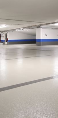 Ein leerer Parkhausbereich mit hellen Wänden und einem grauen Boden. Die Wände sind teilweise mit blauen Streifen dekoriert. Es gibt mehrere Säulen und einige Feuerlöscher an den Wänden.