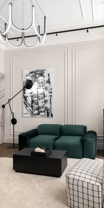 Moderner Sitzbereich im „The Amauris“ Wien mit grünem Samtsofa, hellem Teppich, abstrakter Wandkunst, skulpturaler Stehleuchte, schwarzem Couchtisch, kariertem Sessel und auffälligem, geschwungenen Deckenleuchten-Element.
