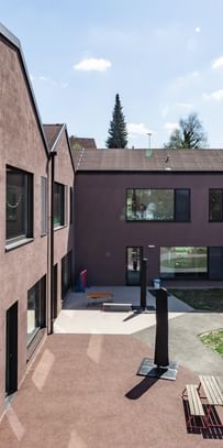 Zweigeschossiges Gebäude mit großen Fenstern und schwarzen Fensterläden. In den oberen Fenstern sind bunte Zeichnungen sichtbar.