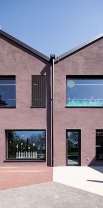 Zweigeschossiges Gebäude mit großen Fenstern. In den oberen Fenstern sind bunte Zeichnungen sichtbar.