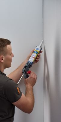 Ein Handwerker, der eine Dichtmasse in die Ecke einer Wand aufträgt. Er trägt ein graues T-Shirt und hält eine Kartusche mit Dichtmittel in der Hand.