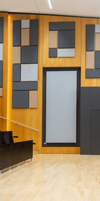 Leerer Hörsaal mit gestuften Sitzreihen, einem Rednerpult mit elektronischer Ausstattung und dekorativen Akustikpaneelen in verschiedenen Formen und Farben an den Holzverkleidungen.