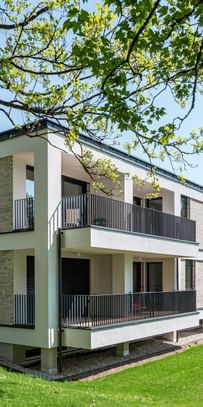 Modernes zweistöckiges Gebäude mit großen Fenstern und Balkonen, gelegen in einer grünen Umgebung mit Bäumen im Hintergrund bei sonnigem Wetter und klarem Himmel.