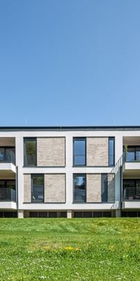 Modernes zweistöckiges Gebäude mit großen Fenstern und Balkonen, gelegen in einer grünen Umgebung mit Bäumen im Hintergrund bei sonnigem Wetter und klarem Himmel.