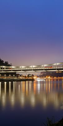 Eine nächtliche Luftaufnahme einer Brücke, die über einen Fluss führt. Die Brücke ist beleuchtet und reflektiert im Wasser.