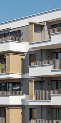 Wohnanlage in der Robert-Stolz-Straße in Linz. Ein modernes, mehrstöckiges Wohngebäude mit hellen Fassaden und zahlreichen Balkonen. Große Fenster sorgen für viel Tageslicht. Vor dem Gebäude stehen Autos auf einer ruhig wirkenden Straße, eingerahmt von gepflegten Grünflächen und Bäumen.