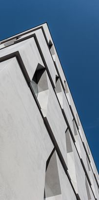 Die Ecke eines Gebäudes, die in die Höhe ragt. Die Fassade ist aus hellem Beton mit schmalen Fenstern, vor einem klaren blauen Himmel.