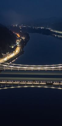 Eine nächtliche Luftaufnahme einer Brücke, die über einen Fluss führt. Die Brücke ist beleuchtet und reflektiert im Wasser.