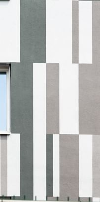 Ein Gebäude mit einer Fassade, die aus vertikalen und horizontalen Streifen in verschiedenen Grautönen und Weiß besteht. In der Mitte der Wand befindet sich ein Fenster mit zwei Fensterscheiben.