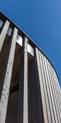 Holzfassade eines Gebäudes von unten. Man blickt auf einen blauen Himmel.