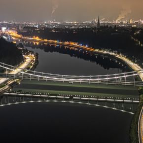 Eine nächtliche Luftaufnahme einer Brücke, die über einen Fluss führt. Die Brücke ist beleuchtet und reflektiert im Wasser.