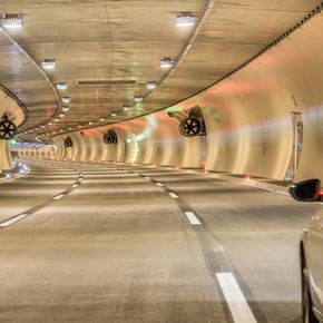 EinTunnel mit sanft geschwungenen Wänden, beleuchtet von bunten Lichtern und auf der rechten Seite ist ein Auto zu sehen, das in den Tunnel einfährt. 