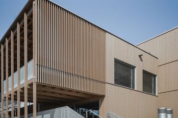Gebäude mit Holzfassade und großen Fenstern. Eine offene Treppe führt zu einem erhöhten Eingangsbereich.