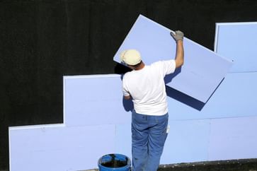 Ein Handwerker trägt eine große, blaue Dämmplatte an eine schwarze Wand.