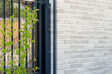 Nahaufnahme einer Gebäudefassade mit hellen Ziegeln, schwarzen Fensterbänken und Metallgittern an den Fenstern. Unten und rechts ist weißer Putz zu sehen, entlang der Wand verläuft ein Streifen mit weißen Kieselsteinen.