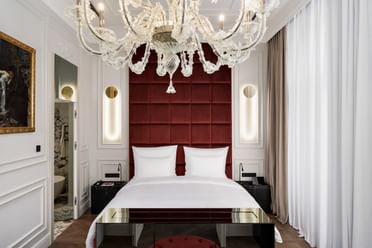 Schlafzimmer der Opera Suite im „The Amauris“ Wien mit großem Doppelbett vor hohem, weinrotem Kopfteil, vertikalen Lichtleisten in weißen Wandpanelen, opulentem Glaslüster und Einblick in ein marmorverkleidetes Badezimmer.