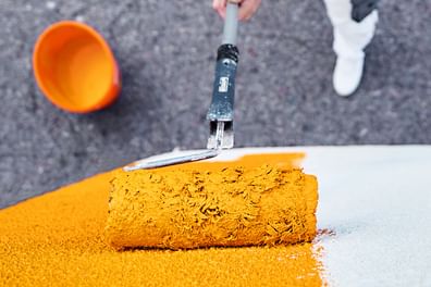 Ein Handwerker in weißer Kleidung trägt mit einem Farbroller orangene Farbe auf eine weiße Wand auf.