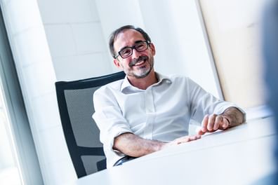 Ein lächelnder Mann mit Brille sitzt an einem Tisch in einem hellen Raum. Er trägt ein weißes Hemd und hat eine entspannte Körperhaltung.