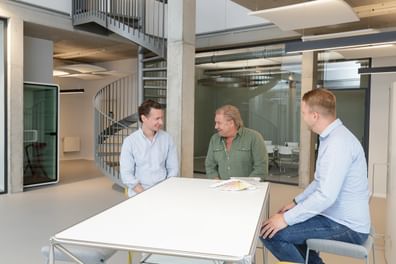 Drei Männer im Gespräch an einem Tisch, Farbtonfächer vor sich, moderne Architektur im Hintergrund