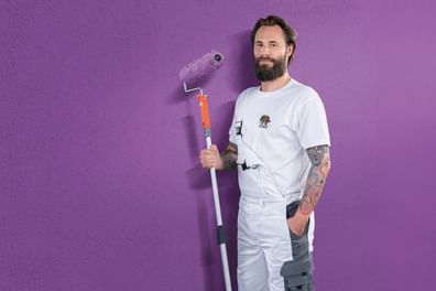 Ein Handwerker in weißer Kleidung steht mit einem Farbroller vor einer lila Wand.
