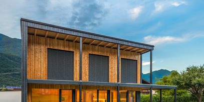 Modernes zweistöckiges Haus mit großen Fenstern und Holzfassade, in der Abenddämmerung beleuchtet. Das Haus ist von grünem Rasen und einem gepflegten Garten umgeben, im Hintergrund sind Berge und weitere Häuser zu sehen.