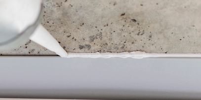 Ein Silikon- oder Dichtmittel wird aus einer Tube auf eine Fuge zwischen einem Betonuntergrund und einer weißen Wandverkleidung aufgetragen.