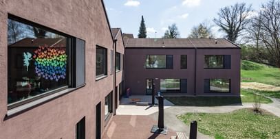 Zweigeschossiges Gebäude mit großen Fenstern und schwarzen Fensterläden. In den oberen Fenstern sind bunte Zeichnungen sichtbar.