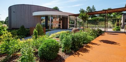  Gebäude mit runder Fassade, umgeben von gepflegten Grünflächen und einem gepflasterten Weg. Im Hintergrund sind Bäume und eine sanfte Hanglage sichtbar.