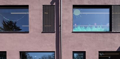 Zweigeschossiges Gebäude mit großen Fenstern. In den oberen Fenstern sind bunte Zeichnungen sichtbar.